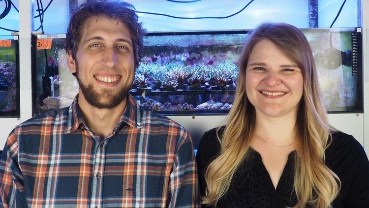 The picture shows Samuel Nietzer and Mareen Möller. They are standing in front of an aquarium with corals in it.