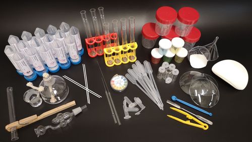 The components of the experimental set are spread out in the picture: Test tubes, spatulas, clamps, a glass spirit burner, chemicals in tubular containers, beakers with lids, etc. 